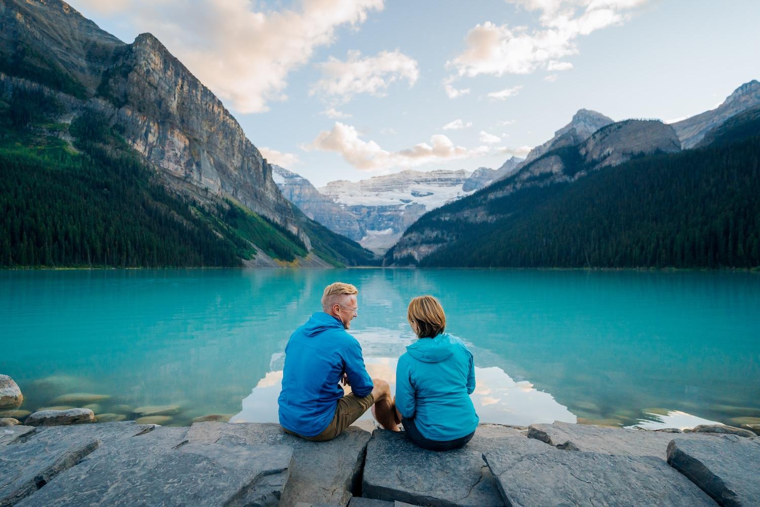 A couple sits on the shore of a turquoise lake in Lake Louise within Banff National Park.