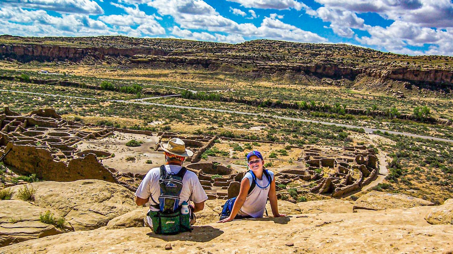 A man sits and a girl is smiling at the camera on the Pueblo Bonito overlook, which displays the full extent of the Pueblo Bonito World Heritage Site below them/Eric Jay Toll