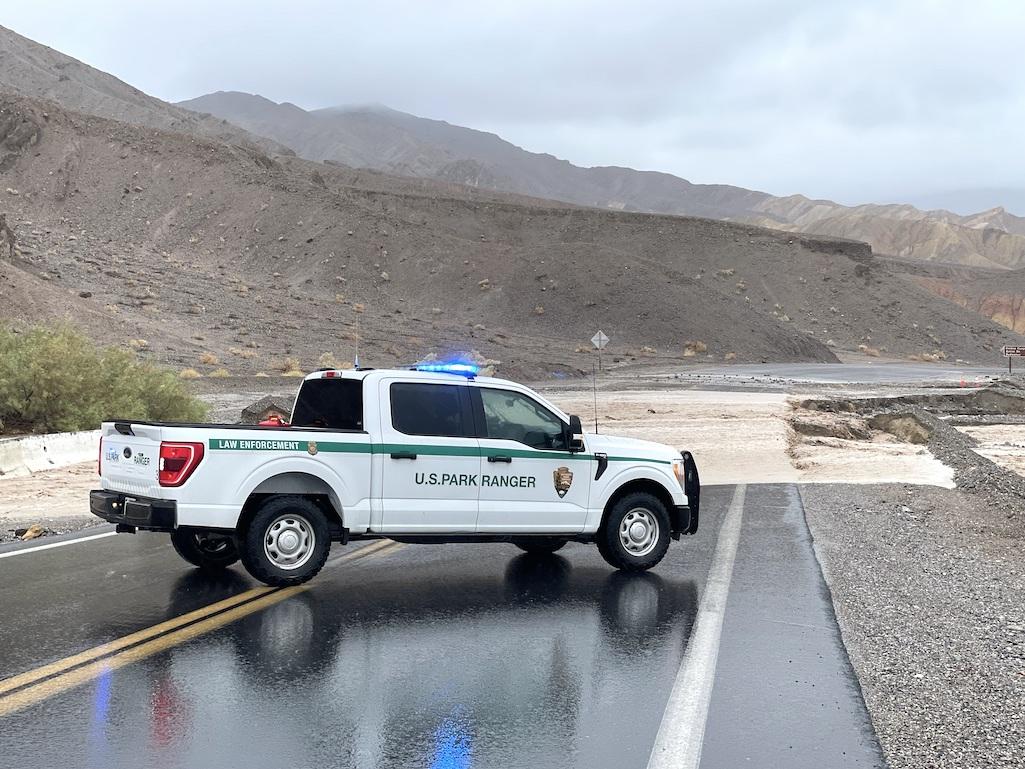 A National Park Service vehicle parked in front of active flooding on Badwater Road, near the junction with CA-190.