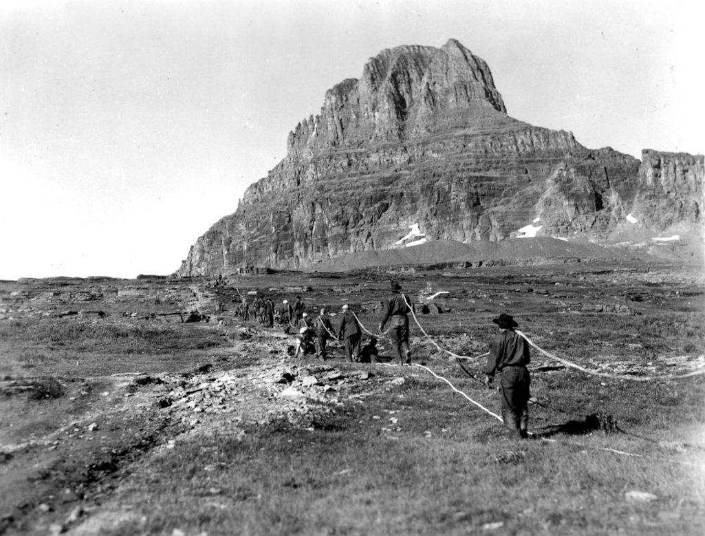 Civilian Conservation Corps crew laying transmountain telephone cable line at Logan Pass, in Hidden Lake area. Shows several crew members forming a continuous chain, each carrying part of the long cable. View is on Logan Pass, looking west toward Clements