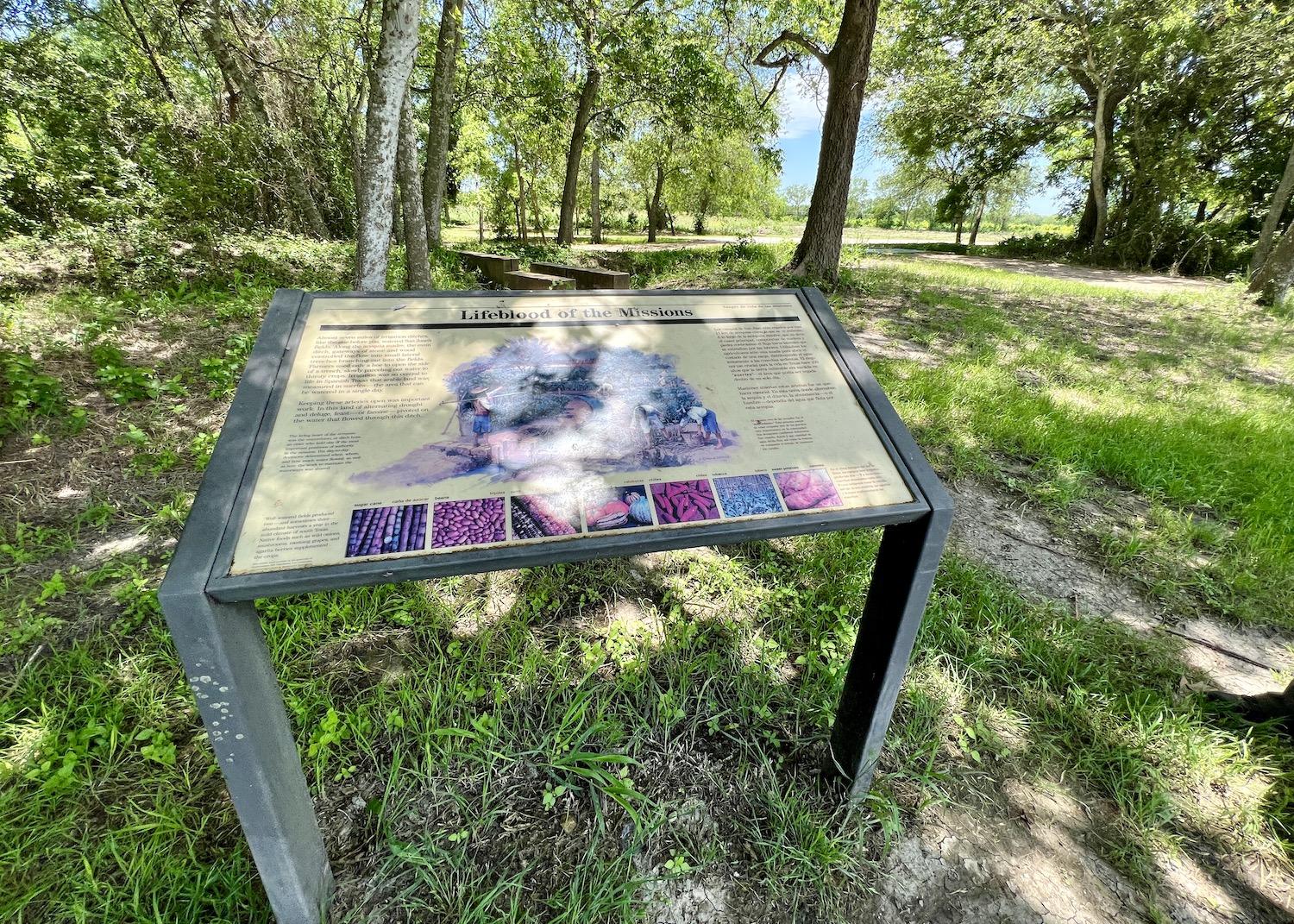 Tucked behind this "Lifeblood of the Missions" sign at Mission San Juan is an acequia and active farm fields.