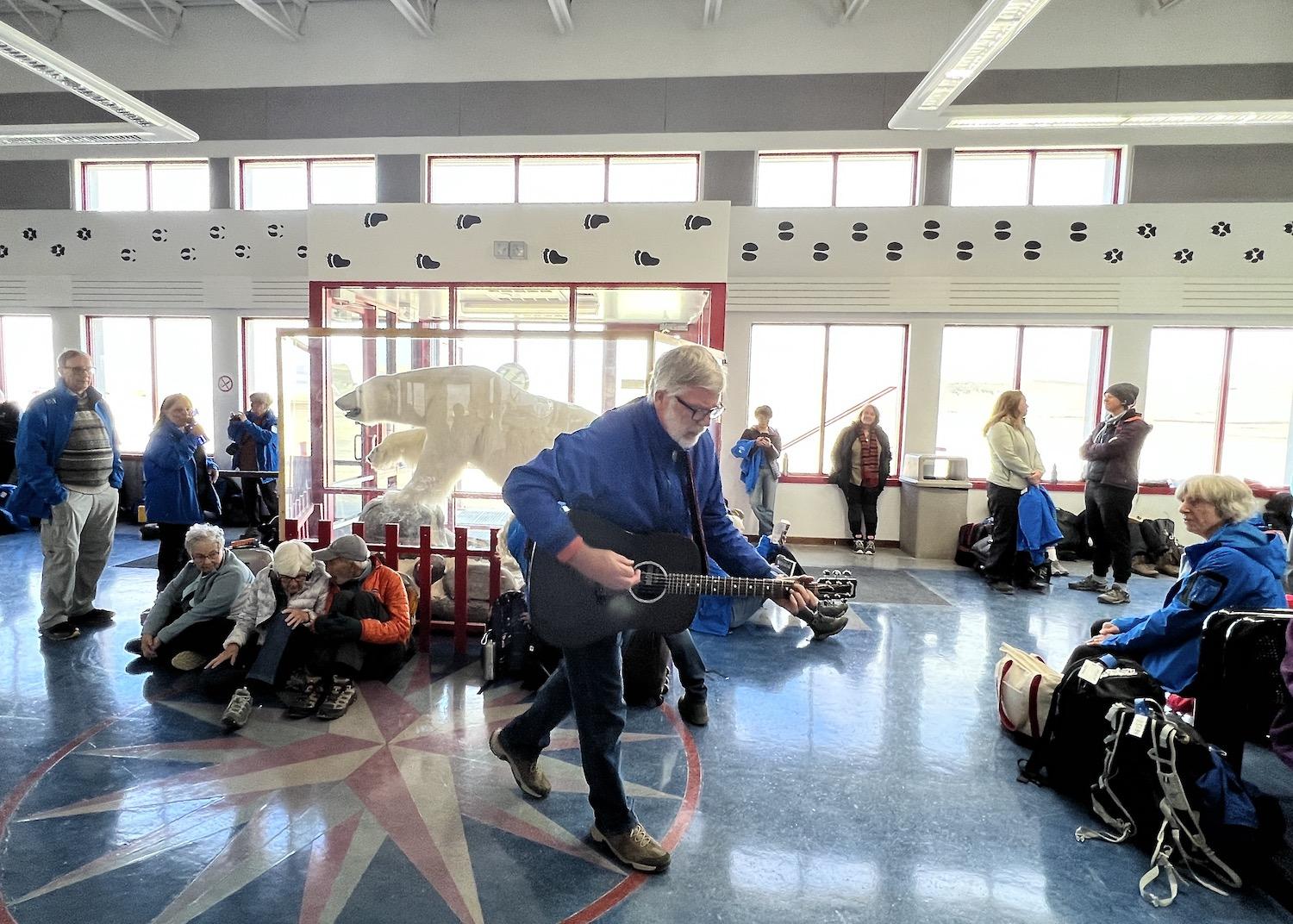At Resolute Bay Airport before a charter flight, Adventure Canada High Arctic Explorer host James (J.R.) Raffan plays for guests in front of a taxidermied polar bear.