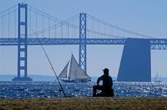 A fisherman sits on a beach with his fishing pole in the sand. A large suspension bridge and sailboats are in the background along the Captain John Smith Chesapeake National Historic Trail.