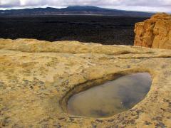 An oval-shaped depression filled with water rests in the surface of a tan cliff. Gray storm clouds fill the sky. Black lava flows below the cliffs appear flat. A distant mountain range lines the edge of the lava flows at El Malpais National Monument