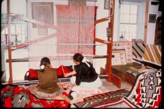 Looking at two Navajo weavers from behind as they kneel in front of a loom at Hubbell Trading Post National Historic Site