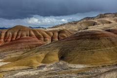A dark, overcast sky and late afternoon sun shining on saturated red-maroon and yellow-green layered hills in the Painted Hills Unit of John Day Fossil Beds National Monument