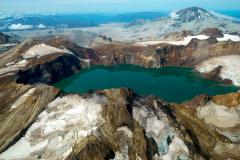 An aerial view of a blue lake within a crater surrounded by rugged mountain and volcano scenery, Katmai National Park and Preserve