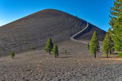 Dark, sandy, barren landscape with a few conifers in the foreground and a 750-foot tall cinder cone with a delineated, curving trail in the background in Lassen Volcanic National Park