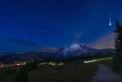 A dark sky during the wee hours of the morning, with the bright flash of a meteor and lights from vehicles, hikers, and the stars at the Sunrise area of Mount Rainier National Park