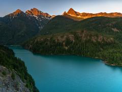 A summer sunrise gilding the tips of the mountains towering over a shaded, teal-blue Diablo Lake at Ross Lake National Recreation Area, a part of the North Cascades National Park Service Complex