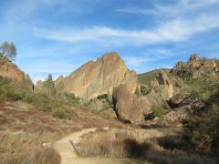 A sandy trail winds through dried grasses and brush and disappears into the rocks and cliffs on the horizon in Pinnacles National Park