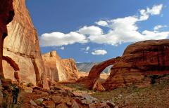A wide-angle view of Rainbow Bridge and surrounding red rock scenery under a blue sky with puffy white clouds at Rainbow Bridge National Monument in Utah