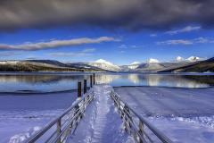 A snow-covered leading line of the dock at Lake McDonald leading the viewer's eye across the water and toward the distant, snow-covered mountains at Glacier National Park
