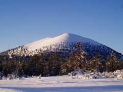 Blue sky and a snowcapped crater towering over a snowy ground at Sunset Crater Volcano National Monument