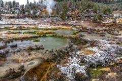 A light dusting of autumn snow over ground with small pools of green, steaming and bubbling hot springs with a boardwalk and steaming Mud Volcano in the background at Yellowstone National Park